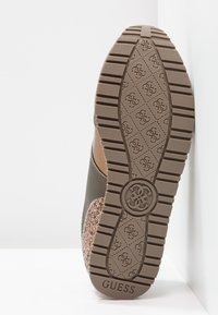 Brauner Gummisohle mit strukturierten Grip-Mustern, die eine Mischung aus glatten und sprenkeligen Oberflächen aufweist. Markenname "GUESS" auf der Ferse geprägt.