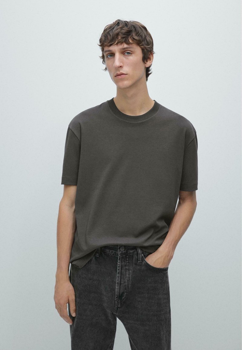 Junger Mann mit kurzen, welligen Haaren, der ein schlichtes dunkelgraues T-Shirt und schwarze Jeans trägt und vor einem hellgrauen Hintergrund steht.