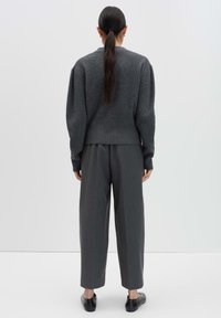 Femme aux longs cheveux noirs en queue de cheval, portant un pull gris foncé, un pantalon gris ample et des chaussures noires à enfiler, se tenant de dos.