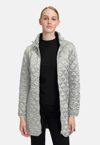 Veste longue matelassée gris clair avec un col haut et une fermeture éclair ; présente un motif en diamant et une texture lisse. Portée sur un haut noir.