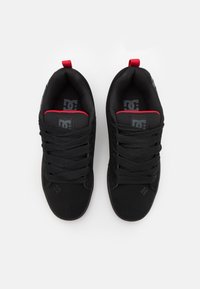 Svarta träningskor med en strukturerad ovandel i tyg, tjocka svarta snören och röda accenter på tungan och hälen. Logotyp synlig på tungan.