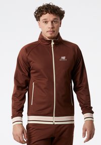 Hnědá sportovní bunda z hladkého materiálu, s předním zipem, bočními kapsami a proužkovanými manžetami v krémové a světle růžové barvě.