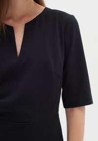 Svart klänning med V-ringning, korta ärmar och smal passform. Tyget har en slät yta med subtila sömmar och en bröstficka.