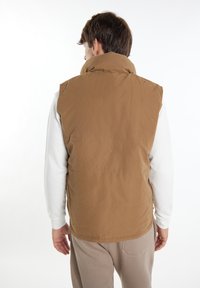 Braune, ärmellose Steppweste mit Kragen, die eine glatte Textur und subtile horizontale Nähte aufweist. Getragen über einem weißen Langarmshirt.