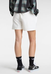 Weiße Shorts mit dezentem Muster, kombiniert mit schwarzen Vans-Sneakern und schwarzen Socken mit weißem Logo. Der Hintergrund ist schlicht.
