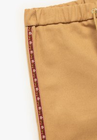Tan joggerpants met een gestructureerde stof, een elastische tailleband en een decoratieve rode bloemenrand aan de zijkant.