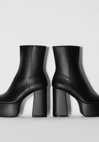 Par de botas negras de cuero con plataforma y tobillo, con gruesos tacones altos, mostradas una al lado de la otra sobre un fondo claro y liso.