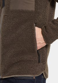 Bruine fleecjas met een textuur van pluizige stof, in contrast met een gladde stof op de borstzak. Met rits gesloten zijzak. Casual ontwerp.