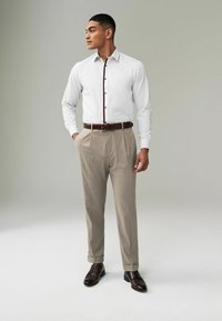 Weißes Langarmhemd mit bordeauxroten Knöpfen, kombiniert mit beigen, gefalteten Hosen und braunen Lederschuhen, vor einem neutralen Hintergrund stehend.
