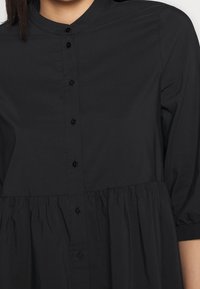 Blusa nera con colletto alto, chiusura a bottoni sul davanti e dettaglio in vita arricciato. Realizzata in un tessuto liscio, presenta maniche corte e bottoni neri.
