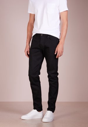 Hombre vestido con jeans negros ajustados, zapatillas blancas y una camiseta blanca, de pie frente a un fondo neutro.