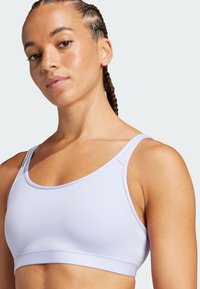 Sostén deportivo de color lila claro con una textura lisa, tirantes anchos y escote redondeado. Presenta costuras a lo largo de los bordes para mayor soporte.