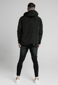 Zwarte hoodie-jack, lichtgewicht materiaal, aansluitend ontwerp. Zwarte jeans met kleurrijk verfspetterspatroon. Witte sneakers.