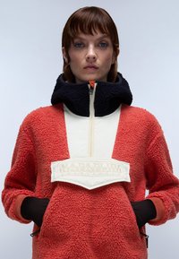 Rød fleece-pullover med en cremefarvet frontpanel, høj krave og sorte detaljer. Har en central logo-patch og sorte lomme-detaljer.