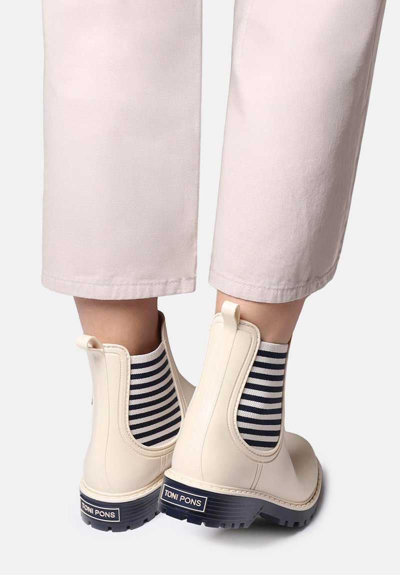 Bottes crème montant à la cheville avec des panneaux latéraux élastiques à rayures noires et blanches, portées avec un pantalon beige clair court, montrant la marque de chaussures "Toni Pons".