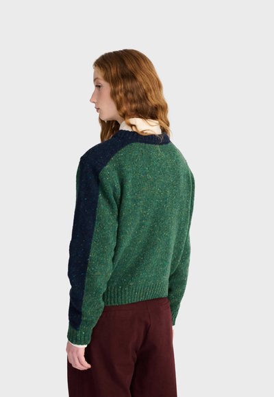 Femme aux cheveux roux ondulés portant un pull texturé vert et bleu marine avec un pantalon marron, de profil sur un fond clair uni.
