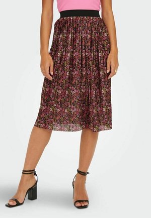 Kvinde iført en plisseret sort floral midi-nederdel med pink og grønne blomster, en pink top og sorte sandaletter med høje hæle.