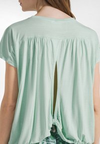 Mintgrøn kortærmet top med slids bagpå og bindedetalje. Blødt, letvægtsstof med rynket tekstur ved skuldrene.