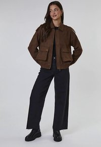 Brązowa zamszowa kurtka bomberka z zamkiem błyskawicznym, dwoma przednimi kieszeniami, ściągaczami przy mankietach oraz pagonami na ramionach, zestawiona z czarnymi spodniami o szerokich nogawkach.