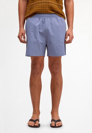 Man draagt lichtblauwe korte broek, bruine textuur shirt en zwarte teenslippers, staand tegen een effen witte achtergrond.