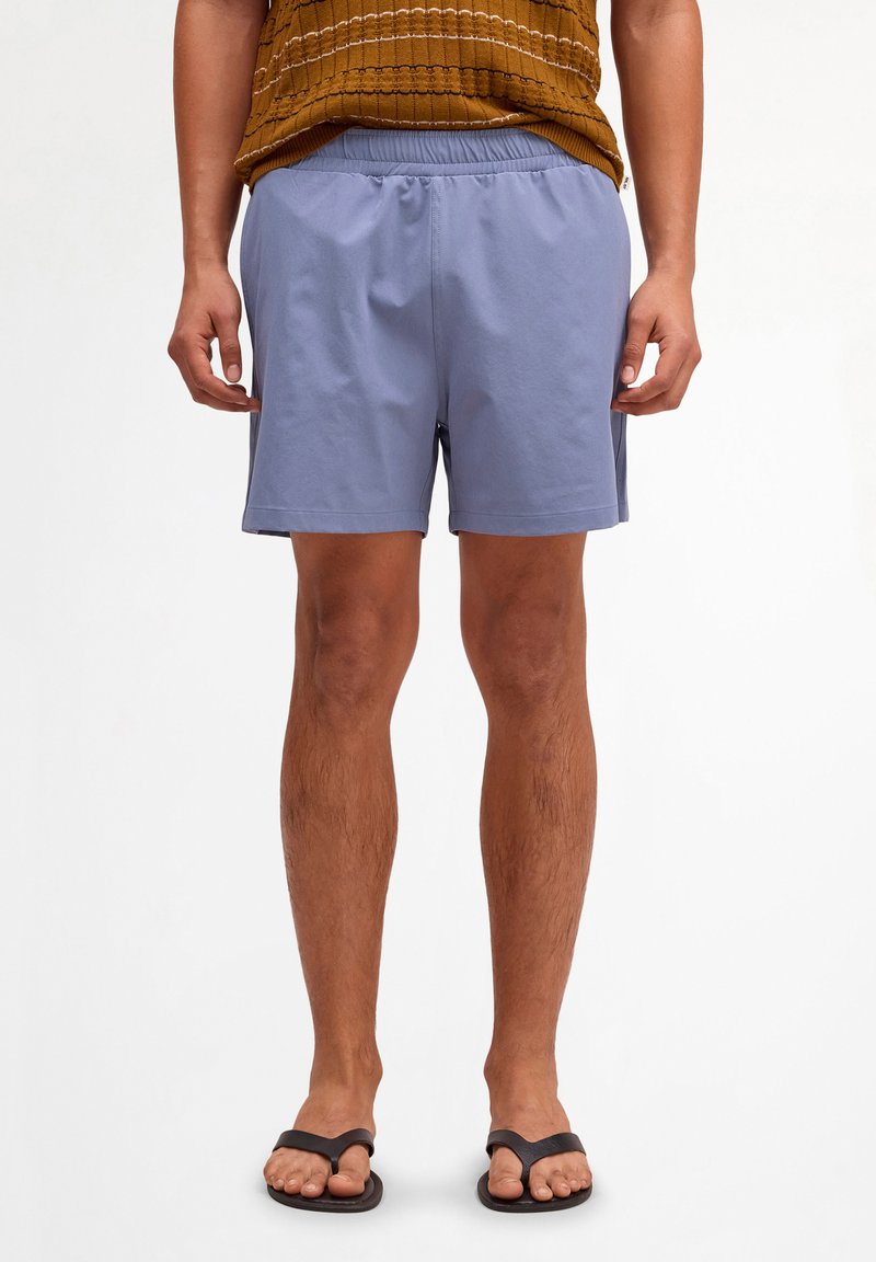 Man wearing light blue shorts, brown textured shirt, and black flip-flops, standing against a plain white background.