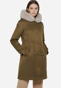 Parka verde oliva con un ampio colletto in pelliccia sintetica, vita regolabile con cordoncino, tasche con bottoni e tessuto liscio. Finiture in metallo dorato.