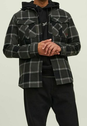 Camisa de cuadros en negro y gris con dos bolsillos en el pecho, cierre de botones y una textura suave, llevada sobre un sudadera con capucha negra.
