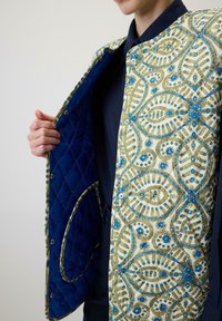 Gequilted jack met een sierlijke crèmekleurige stof met groene en blauwe bloemmotieven, op drukknopen en een marineblauwe voering.