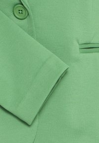 Groene blazer met textuur, mouw gekruist over de voorkant van het jasje, met bijpassende groene knoop en zakdetail.