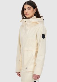 Helle beige wasserdichte Jacke mit verstellbarem Kapuzen, Frontreißverschluss, seitlichen Taschen und schwarzem runder Logo-Patch auf dem linken Ärmel.