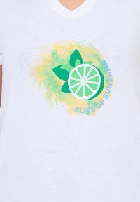Camiseta blanca de cuello en V con un gráfico de lima verde, hojas verdes y el texto "UN TROZO DE SOL", acentuada por un fondo de salpicaduras de acuarela amarilla.
