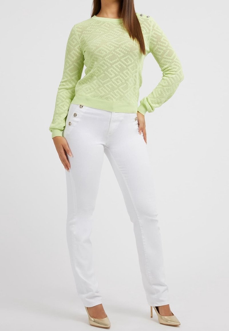 Femme portant un pull à manches longues texturé vert clair et un pantalon blanc avec des boutons dorés, assortis à des chaussures à talons hauts dorées, debout.