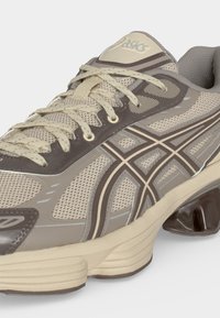 Μπεζ και καφέ παπούτσι για τρέξιμο Asics με επάνω μέρος από δίχτυ, σχεδιασμένες πλαϊνές ρίγες, αντικραδασμική σόλα και μπεζ κορδόνια δεμένα σε λευκό φόντο.