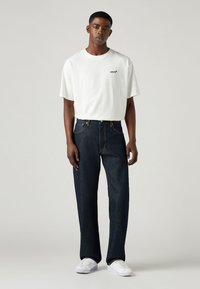 Camiseta blanca de manga corta con el logo de "Levi's", combinada con jeans rectos de pierna oscura y zapatillas blancas. Diseño minimalista, ajuste casual.