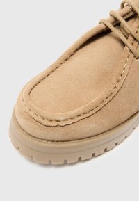 Chaussure en suede beige avec un bouts rond, des accents cousus et une semelle en caoutchouc dotée d'un motif de semelle. Les lacets sont positionnés au centre.