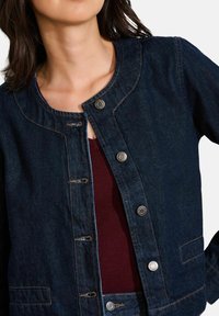 Veste en denim bleu foncé avec un col rond, fermeture à boutons sur le devant et couture contrastante. Présente une texture lisse et deux poches avant.