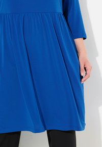 Niebieska, do kolan sukienka wykonana z gładkiego, elastycznego materiału. Posiada marszczoną talię i rękawy o długości 3/4, zestawiona z czarnymi spodniami.