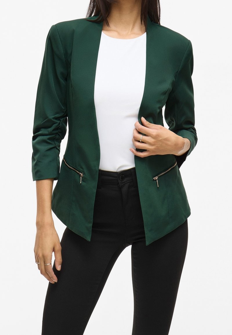 Femme portant un blazer vert foncé avec des poches zippées par-dessus un haut blanc et un pantalon noir, tenant le revers du blazer d'une main.