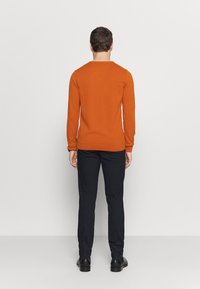 Oranssi neulottu pusero pyöreällä kaula-aukolla, resorit hihansuissa ja helmassa. Päällä tummat housut ja mustat kengät. Takakuvassa, yksivärinen tausta.