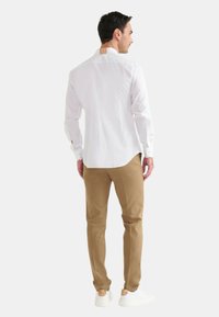 Uomo con una camicia bianca a maniche lunghe, pantaloni beige e scarpe da ginnastica bianche, in piedi di spalle su uno sfondo chiaro e uniforme.