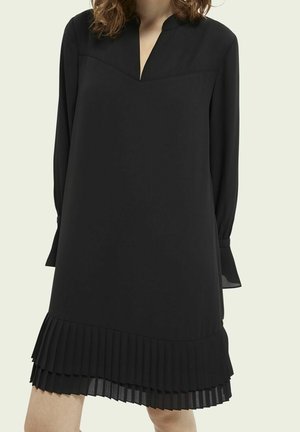 Femme portant une robe noire à manches longues avec un décolleté en V et un ourlet plissé, sur un fond clair uni.