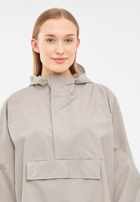 Anorak ligero en gris claro, con capucha, bolsillo frontal y mangas anchas. Textura suave con un acabado mate y diseño minimalista.