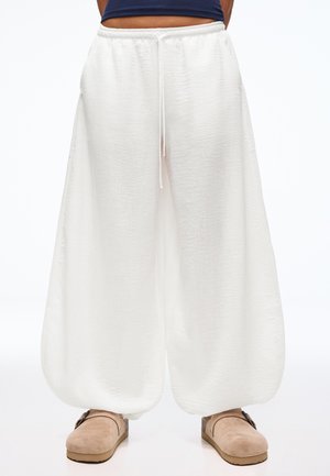Luźne białe spodnie o fakturze, z elastycznym paskiem i sznurkiem, zestawione z beżowymi wsuwanymi butami i granatowym krótkim topem.