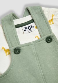 Zaļš bērnu kombinezons ar pogājām plecu siksnām, slāņots pār baltu bodiju ar dzelteniem žirafu rakstiem, izmēra birka norāda 3-6 mēnešus.