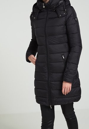 Femme portant un long manteau matelassé noir avec fermeture éclair à l’avant, poches zippées et un col montant sur un fond clair uni.
