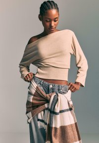 Beige off-shoulder cropped sweater, ribbed hem, paired with light denim jeans, cinched at the waist with a patterned scarf in cream and brown.