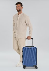 Valigia rigida di colore blu navy lucido con motivo a chevron, manico retrattile e quattro ruote per la mobilità. Include una maniglia laterale.