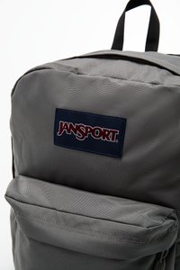 Sac à dos gris Jansport avec une texture de tissu lisse, poche avant zippée et un badge logo bleu marine. Design compact et rectangulaire.