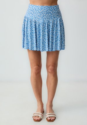 Jupe mini bleue à fleurs avec un design plissé, présentant des fleurs blanches sur un fond bleu clair, fabriquée en tissu doux avec une taille élastique.