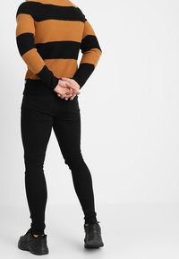 Neulottu pusero, jossa on vaakasuorat sinapinväriset ja mustat raidat, yhdistetty kapealahkeisiin mustiin farkkuihin ja mustiin lenkkareihin. Takakatsaus, kädet ristissä selän takana.